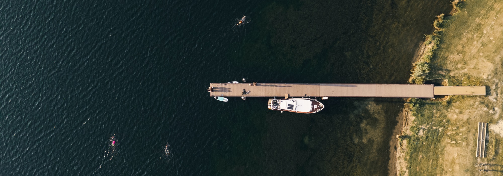 Luchtfoto van een lange steiger aan de Maasplassen, met een boot, suppers en zwemmers in het heldere water.