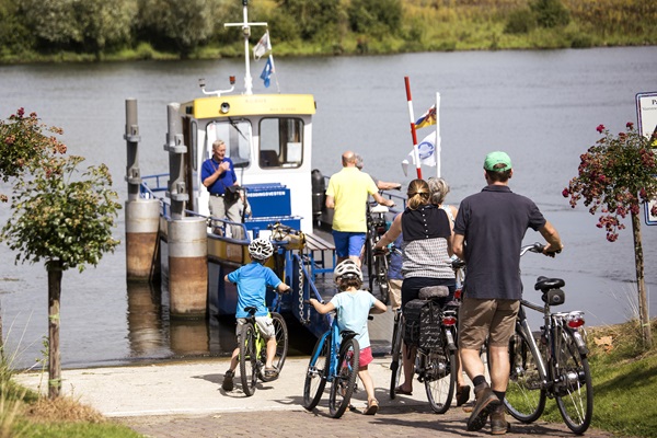 Fietsers, waaronder kinderen en volwassenen, stappen in Blitterswijck met hun fietsen op een veerpont over de Maas op een zonnige dag.