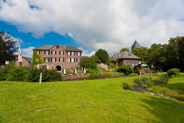 Blick auf den historischen Ortskern von Brüggen mit Schloss, Wassermühle und Uferterrasse, umgeben von viel Grün und klarem Himmel.