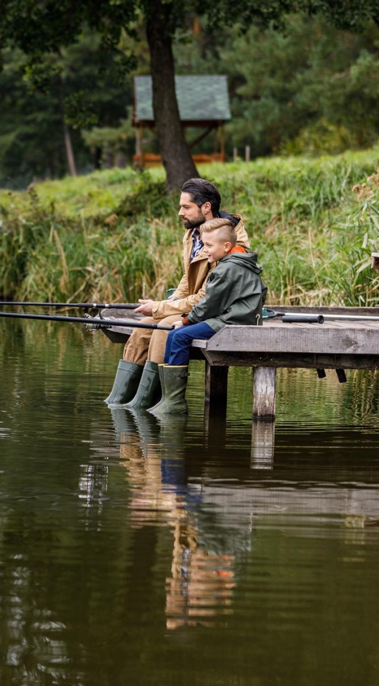 Vader en zoon zitten samen te vissen aan de rand van een visvijver in Limburg, omringd door groene natuur.