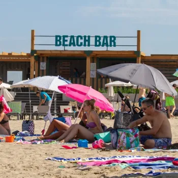 Mensen genieten van een zonnige dag op het strand bij de Maasplassen, met parasols en een beach bar op de achtergrond.