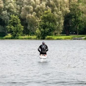 Persoon op een fliteboard vaart over rustig water met op de achtergrond een groene oever vol bomen.