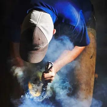 Hoefsmid brandt een hoefijzer op de hoef van een paard tijdens het beslaan, met rookwolk in beeld.