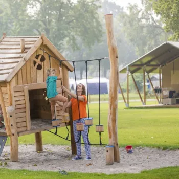 Moeder helpt haar kind op een houten speeltoestel bij een glamping op recreatiepark De Leistert, met glampingtenten op de achtergrond.